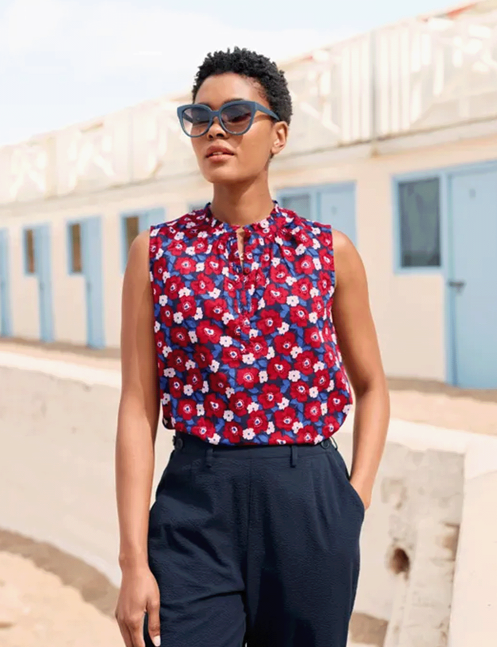 Woman standing in front of beach huts wearing the Flower Fields Sleeveless Top with navy trousers